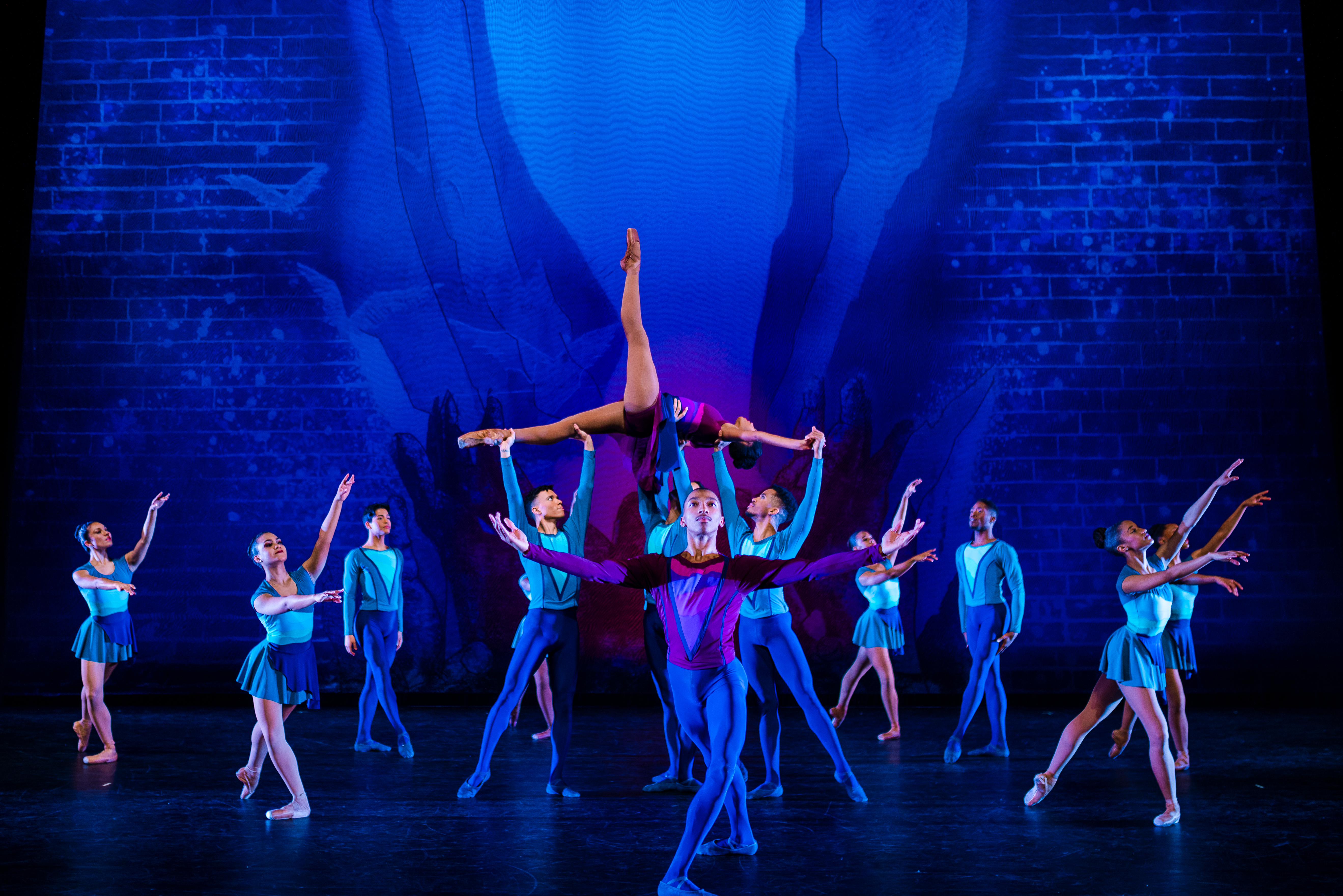 Male and female ballet dancers in blue and purple costumes, dancing against blue background, with one female ballet dancer being lifted.